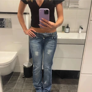 Lågmidjade bootcut jeans i ljusblå denim - Säljer ett par riktigt snygga lågmidjade jeans i ljusblå denim med bootcut passform. Jeansen har slitna detaljer framtill, klassiska fickor och dragkedja. Perfekta för dig som gillar 00-talsvibbar och vill ha en avslappnad men trendig look.