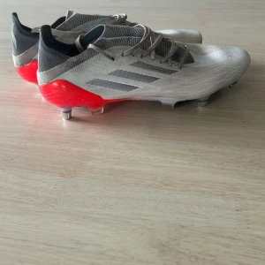 ADIDAS X SPEEDFLOW .1 SG - Säljer ett par Adidas fotbollsskor med snygg design i vitt och rött. Skorna har metall-dobbar, snörning och en strömlinjeformad siluett med mesh-överdel för bra ventilation. De har svarta och grå detaljer samt klassiska Adidas-ränder på sidan. Perfekt för dig som vill ha grymt grepp på planen.