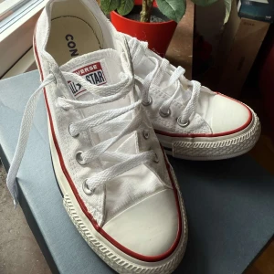 Converse All Star vita sneakers - Säljer ett par klassiska Converse All Star sneakers i vitt med röda och svarta detaljer runt sulan. Skorna har snörning, rund tå och platt sula. Tillverkade i bomullstyg med gummisula. Perfekta för dig som gillar ikonisk streetstil och vill ha ett par schyssta låga sneakers.