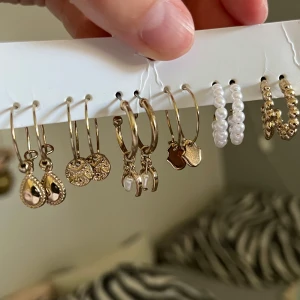 Set med 6 par örhängen i guld och pärlor - Säljer ett snyggt set med 6 par örhängen i olika stilar. Du får guldiga hoops med droppformade hängen, runda medaljonger, små pärlor, hjärtformade berlocker, samt två par med vita pärlor och guldiga kulor. Perfekt för att mixa och matcha till din egen vibe.