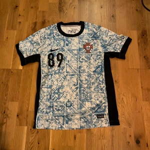 Portugal Nike fotbollströja blåvit - Säljer en Portugal fotbollströja från Nike med unikt blåvitt mönster och svarta detaljer. Tröjan har korta ärmar, tryckt nummer 89 på bröstet och broderat Portugal-emblem. Tillverkad i lätt och ventilerande material, perfekt för match eller träning. Som har namnet AGATA på ryggen.