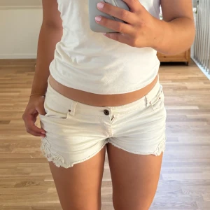 Vita jeansshorts med spetsdetalj - Snygga vita jeansshorts med låg midja och fina spetsdetaljer längs bensluten. Klassisk femficksmodell med bälteshällor och knappstängning. Perfekta för varma sommardagar och ger en fräsch look till din outfit.