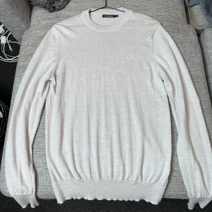 Ljusbeige merinoulltröja J.Lindeberg - Snygg ljusbeige rundhalsad tröja från J.Lindeberg i mjuk merinoull. Klassisk och stilren design med diskret logga på bröstet, ribbade muddar och lång ärm. Perfekt för dig som gillar cleana och tidlösa plagg med premiumkänsla. Loggan på bild 3 sitter löst men inget som syns under användning, går att sy ditt om man vill.