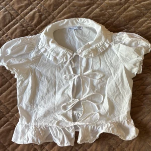 Vit blus med snörning och volang - Supersöt vit blus från Bershka med puffiga korta ärmar, volangkant och snörning framtill. Den har en rund krage med volangdetaljer och flera rosetter längs öppningen. Perfekt för dig som gillar romantisk och lekfull stil.