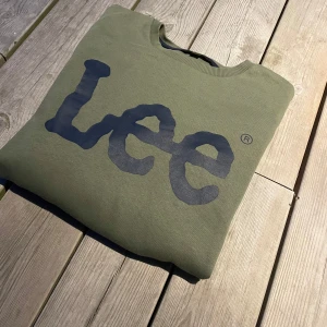 Olivgrön Lee sweatshirt med tryck - Snygg olivgrön sweatshirt från Lee med stort svart logotryck på bröstet. Tröjan har rund hals, långa ärmar och ribbade muddar vid ärmslut och nederkant. Perfekt för chill dagar och enkel att matcha med jeans eller joggers. Axelbredd: 41cm  Längd: 65cm
