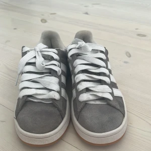 Adidas Campus gråa - Säljer ett par Adidas Campus sneakers i grå mocka med vita detaljer och klassiska tre ränder på sidan. Skorna har vit sula med gummidetalj och rund tå. Tjocka vita snören och Adidas-logga på hälen. Perfekt för dig som gillar retrostil och streetwear.De har en lite gel fläck som de har haft sen jag köpte dom.