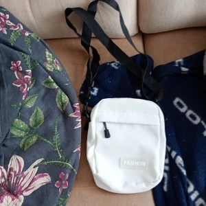 Vit axelväska med svart rem - Snygg vit axelväska från Fashion med svart justerbar rem och praktisk dragkedja. Perfekt storlek för mobilen och småsaker, stilren design med ett litet märke framtill. Enkel att matcha med olika outfits och passar dig som gillar minimalistisk stil.