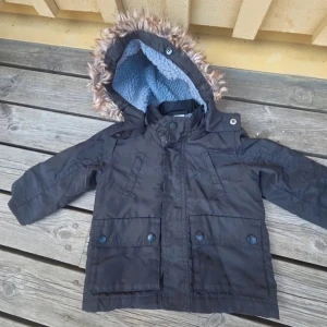 Svart parkajacka med fuskpäls - Svart parkajacka med mysig blå teddyfoder på insidan och huva med fuskpäls. Jackan har stora fickor med knappar framtill och en patch på ena ärmen. Perfekt för kalla dagar och har en klassisk, stilren look. 