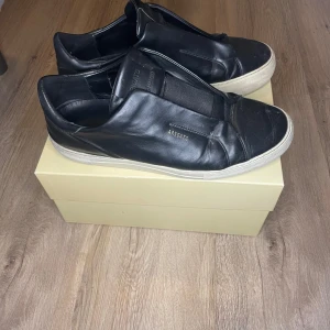 Svarta sneakers från Axel Arigato - Snygga svarta sneakers från Axel Arigato med minimalistisk design och diskret logga i guld på sidan. Skorna har en rund tå, platt vit sula och är tillverkade i slätt skinn. Perfekta för dig som gillar stilrena och cleana sneakers.