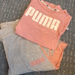Grå sett från Puma med rosa och grå detaljer - Snygga grå sett Puma med rosa sidopaneler och logga på benet. O en rosa t-shirt  Byxorna o t-shirt  har en avslappnad passform och är gjorda i mjukt bomullsmaterial, perfekta för chill eller träning. Fräscha färger och sportig vibe.