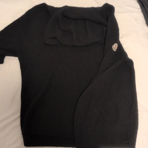 Svart stickad hoodie från Moncler - Svart stickad hoodie från Moncler med klassisk logga på ärmen. Tröjan har huva med snörning och är långärmad. Grovstickad struktur ger en cool och avslappnad vibe, perfekt för dig som gillar streetwear och exklusiva märken.