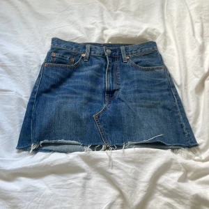 Levi's jeanskjol med rå kant, blå - Säljer en blå jeanskjol från Levi's i klassisk modell med fem fickor och rå, fransig kant nedtill. Kjolen är kort och har normal passform, perfekt för dig som gillar en chill och avslappnad stil. Ikonisk Levi's-lapp baktill och snygga detaljer.