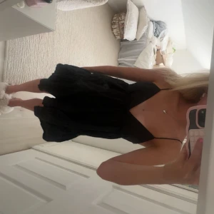 Svart kort klänning med snörning - Säljer en svart kort klänning med v-ringad front och tunna axelband. Klänningen har volang i nederdelen och snörning i ryggen för en snygg detalj. Materialet känns lätt och passar perfekt för varmare dagar. Enkel och stilren design som är lätt att matcha.