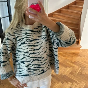 Beige och mörkgrön stickad tröja - Säljer en fluffig stickad tröja med beige botten och mörkgröna zebraränder. Tröjan har en loose passform, rund hals och långa ärmar. Perfekt för dig som gillar djurmönster och vill ha något riktigt mysigt och coolt i garderoben.