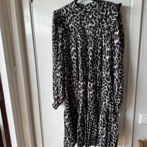 Leopardmönstrad klänning Pernilla Wahlgren XS - Snygg kort klänning från Pernilla Wahlgren i leopardmönster med svart och beige ton. Modellen har lång ärm, volangdetaljer vid axlarna och en liten guldig knapp framtill. Materialet är mjukt och känns som bomull. Perfekt för dig som vill sticka ut med coola detaljer.