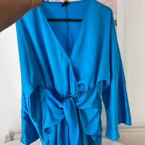 Blå omlottklänning från Gina Tricot - Säljer en snygg kort blå omlottklänning från Gina Tricot med breda ärmar och stor knytrosett i midjan. Klänningen har v-ringad hals och är gjord i ett glansigt, mjukt material som ger en lyxig känsla. Perfekt för dig som vill sticka ut med färg!