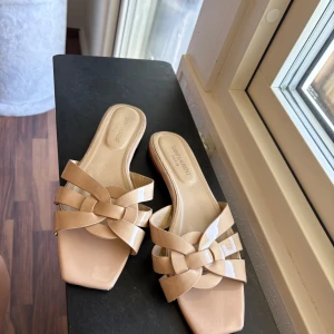 Beige sandaler från Saint Laurent - Snygga beige sandaler från Saint Laurent med flätad design över foten och fyrkantig tå. Skorna är tillverkade i blankt skinn och har en platt sula som gör dem bekväma att bära. Perfekta för dig som gillar stilrena och lyxiga detaljer.