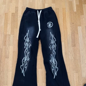Hellstar mjukisbyxor - Svarta mjukisbyxor med vita flames längs benen och en vit logga på höften. Byxorna har breda ben, vit snörning i midjan och är gjorda i mjukt bomullsmaterial. Perfekt för chill eller streetwear-stil.