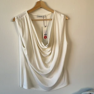 Vit draperad linnetopp från Kappahl - Snygg vit linnetopp från Kappahl i storlek L med draperad front och mjukt fall. Toppen är ärmlös och har en cool omlottkänsla framtill. Materialet är lent och har en lätt glans, perfekt för att lyfta din look med en clean vibe.