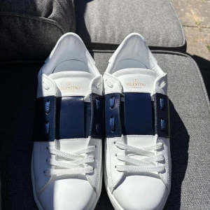 Valentino skor - Snygga sneakers från Valentino Garavani i vitt skinn med bred marinblå panel över mitten och matchande marinblå snörning. Låg modell med rund tå och platt sula med coola nitar längs kanten. Perfekt för dig som gillar stilrena och lyxiga detaljer STORLEK 43 men passar 42