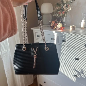 Svart axelväska från Valentino - Snygg svart axelväska från Valentino med silverkedjor som axelremmar och ett V-emblem framtill. Väskan har en stilren, rektangulär form och är tillverkad i glansigt skinn. Perfekt för dig som gillar en lyxig och modern look.