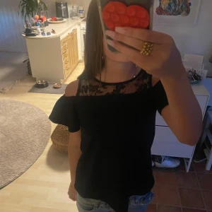 Svart topp med spets och öppna axlar - Säljer en svart festtopp med snygga öppna axlar och spetsdetaljer upptill. Blusen har korta ärmar med volang och ett transparent spetsparti med spets mönster vid halsen.