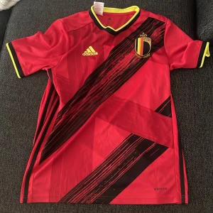 Belgien fotbollströja Adidas röd - Säljer en officiell Belgien fotbollströja från Adidas i röd polyester med svarta och gula detaljer. Tröjan har korta ärmar, V-ringad krage med gul kant och ett coolt diagonalt svart mönster framtill. Belgiska landslagets emblem på bröstet och Adidas-logga i gult.