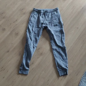 Grå mjukisbyxor från Hoodrich M - Säljer ett par grå mjukisbyxor från Hoodrich i storlek medium. Byxorna har snörning i midjan, ribbade muddar vid bensluten och Hoodrich-logga tryckt på benet. Sköna och stilrena, perfekta för chill eller träning.