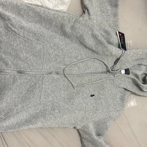 Grå hoodie från Polo Ralph Lauren - Snygg grå hoodie från Polo Ralph Lauren i storlek S. Tröjan har dragkedja, huva med snörning och ett mörkblått Polo-logo broderat på bröstet. Tillverkad i mjuk bomullsmix, perfekt för chill dagar. Klassisk passform och ribbade muddar vid ärmar och nederkant.