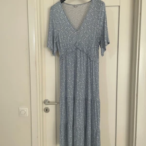 Blommig blå långklänning Happy Holly - Supersöt långklänning från Happy Holly i ljusblått med små vita och blå blommor. Klänningen har v-ringning, korta ärmar och ett mjukt fall. Tillverkad i skön polyester, perfekt för vår och sommar. Blommigt mönster ger en fräsch och romantisk vibe.