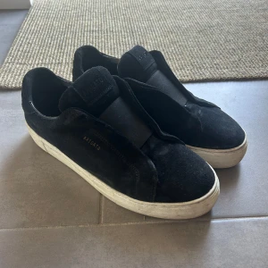 Svarta Arigato sneakers i mocka - Svarta sneakers från Axel Arigato med stilren design och bred elastisk rem över vristen. Ovandel i mjuk mocka och insida i skinn, med vit platt gummisula. Diskret Arigato-logga i guld på sidan. Perfekt för dig som gillar minimalistisk streetstil.