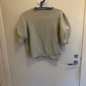 Grågrön stickad tröja med puffärm - Säljer en grågrön stickad tröja med puffiga, korta ärmar och rund hals. Tröjan har ribbade muddar vid ärmslut och nederkant, vilket ger en snygg och chill look. Perfekt för dig som gillar enkel stil med en twist.