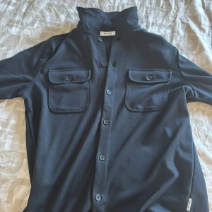Mörkblå overshirt från Once & Sons - Snygg mörkblå overshirt med hög krage och två stora bröstfickor med knappar. Långärmad modell med knäppning hela vägen framtill. Materialet känns mjukt och har en lätt struktur. Perfekt att bära som lager över t-shirt eller hoodie.