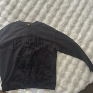 Svart sweatshirt från Lee Jeans - Svart sweatshirt från Lee Jeans med stor, ton-i-ton logga och emblem på bröstet. Tröjan har rund hals, långa ärmar och ribbade muddar vid ärmslut och nederkant. Perfekt för chill dagar och enkel att matcha med jeans eller joggers.