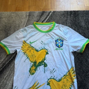  Brasilien fotbollströja  - Säljer en vit Brasilien fotbollströja från Nike med gröna och gula detaljer. Tröjan har korta ärmar, grönt och gult runt kragen och ärmsluten samt stora gula fåglar tryckta på framsidan. Brasilien-emblem och Nike-logga på bröstet.