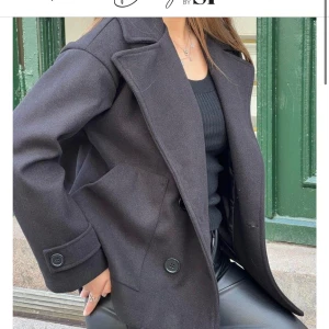 Svart kappa från Design by Si - Stilren svart kappa med bred krage och stora fickor från Design by SP. Jackan har dubbelknäppning med stora svarta knappar och är helfodrad. 