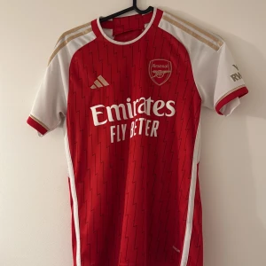Arsenal hemmatröja 2023/24 - Röd och vit Arsenal fotbollströja från Adidas med gulddetaljer på axlarna och klubbmärke på bröstet. Tröjan har korta ärmar, rund hals och ett subtilt blixtmönster över hela framsidan. Emirates Fly Better-logga tryckt på bröstet. 100% original köpt från Adidas butik i Östterike