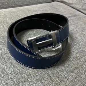 Mörkblått skärp med silverfärgat H-spänne - Snyggt mörkblått bälte med ett stort H-format spänne i silver. Bältet har vita sömmar längs kanterna och är gjort i ett slätt material som liknar skinn. Perfekt accessoar för att lyfta din outfit.