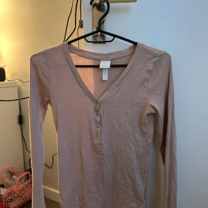 Beige ribbad långärmad topp med knappar - Säljer en beige ribbad långärmad topp från H&M med v-ringning och knappar framtill. Perfekt för lager-på-lager och enkel att matcha med jeans eller kjol. Snygg och stilren modell med smal passform.