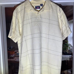 Gul Piké från Timber Creek by Wrangler - Säljer en ljusgul piké från Timber Creek by Wrangler. Tröjan har ett rutigt mönster i svart och klassisk krage. Tillverkad i en blandning av bomull och polyester, perfekt för varma dagar.