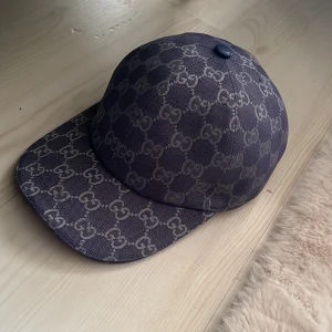 Gucci keps med GG-mönster - Säljer en mörkblå keps från Gucci med klassiskt GG-mönster i grått. Kepsen har böjd skärm och justerbar rem baktill. Tillverkad i bomullsmix med tydlig logotypdetalj. Perfekt för dig som gillar exklusiva accessoarer, Skick 10/10 knappt använd.