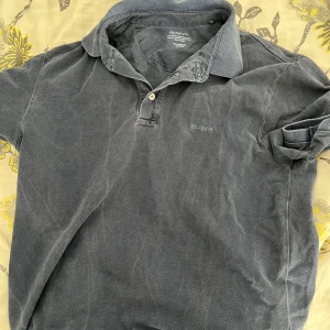 Mörkblå pikétröja från GANT - Säljer en klassisk mörkblå pikétröja från GANT i storlek medium. Tröjan har korta ärmar, krage och knappar framtill. Perfekt för en avslappnad men stilren look. GANT-logga broderad på bröstet.