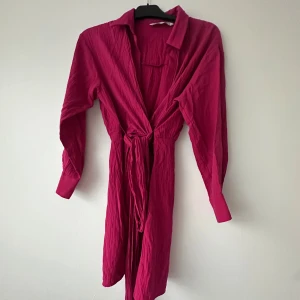 Rosa långärmad klänning med knytband - Säljer en snygg rosa klänning med lång ärm och krage. Klänningen har knytband i midjan för en justerbar passform och är tillverkad i ett lätt, strukturerat material. Perfekt för dig som vill ha en färgstark look!