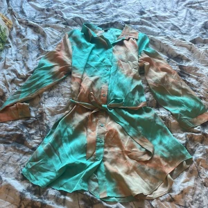 Turkos och beige skjortklänning med bälte - Säljer en snygg skjortklänning i turkos och beige med knappar framtill och matchande bälte i midjan. Klänningen har långa ärmar och är tillverkad i ett lätt, glansigt material som känns lyxigt. Perfekt för dig som vill sticka ut med färg!