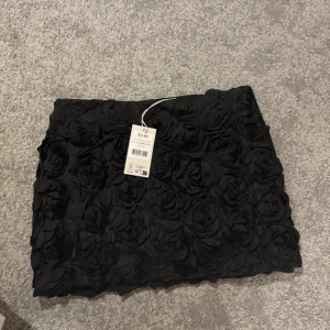 Svart kjol med 3D-blommor - Supersnygg svart kortkjol med 3D-blommor över hela tyget. Kjolen har en tight passform och är perfekt för dig som vill sticka ut. Blommorna ger en cool och unik look. Passar till många olika stilar och är lätt att matcha med olika toppar. Storlek 40