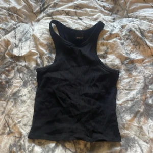 Svart ribbad linnetopp - Säljer en svart ribbad linnetopp från Gina. Toppen har en sportig skärning med breda axelband och ärmlös design. Perfekt att matcha med jeans eller kjol för en enkel och stilren look.