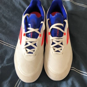 Vita Adidas fotbollsskor med röda och blå detaljer - Helt nytt.Säljer ett par vita Adidas fotbollsskor med röda ränder på sidorna och blå insida. Skorna har snörning och en sportig design med rund tå och platt sula. Perfekta för dig som vill sticka ut på planen!
