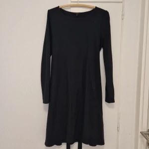 Mörkblå långärmad klänning från COS - En stilren mörkblå klänning från COS med lång ärm och rund halsringning. Klänningen har ett knytband i midjan för en snygg siluett och är tillverkad i ett mjukt material. Perfekt för dig som gillar minimalistisk design. Framsidan är i annat material än övriga klänningen.