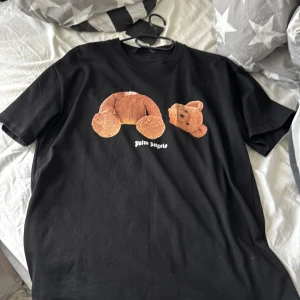 Svart t-shirt med nalletryck från Palm Angels - Svart t-shirt från Palm Angels med ett stort tryck av en brun nalle på framsidan och vit Palm Angels-logga på ryggen. Klassisk passform och rund halsringning. Perfekt statement-plagg för dig som gillar streetwear.