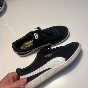 Svarta Puma sneakers med vit sula och logga - Säljer ett par klassiska svarta Puma sneakers med vit sula och vit sidodetalj. Skorna har snörning och rund tå, samt Puma-logga i guld på hälen och plösen. Perfekta för en stilren och sportig look.